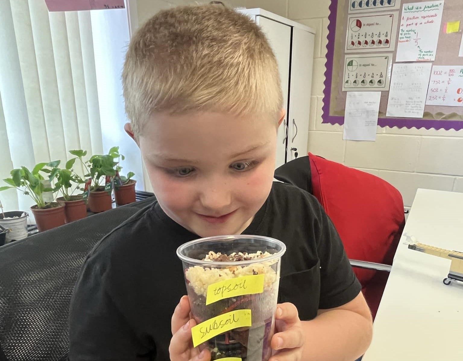 A young blonde boy looking eagerly into a cup of different soil examples. In the background is a classroom setting.  